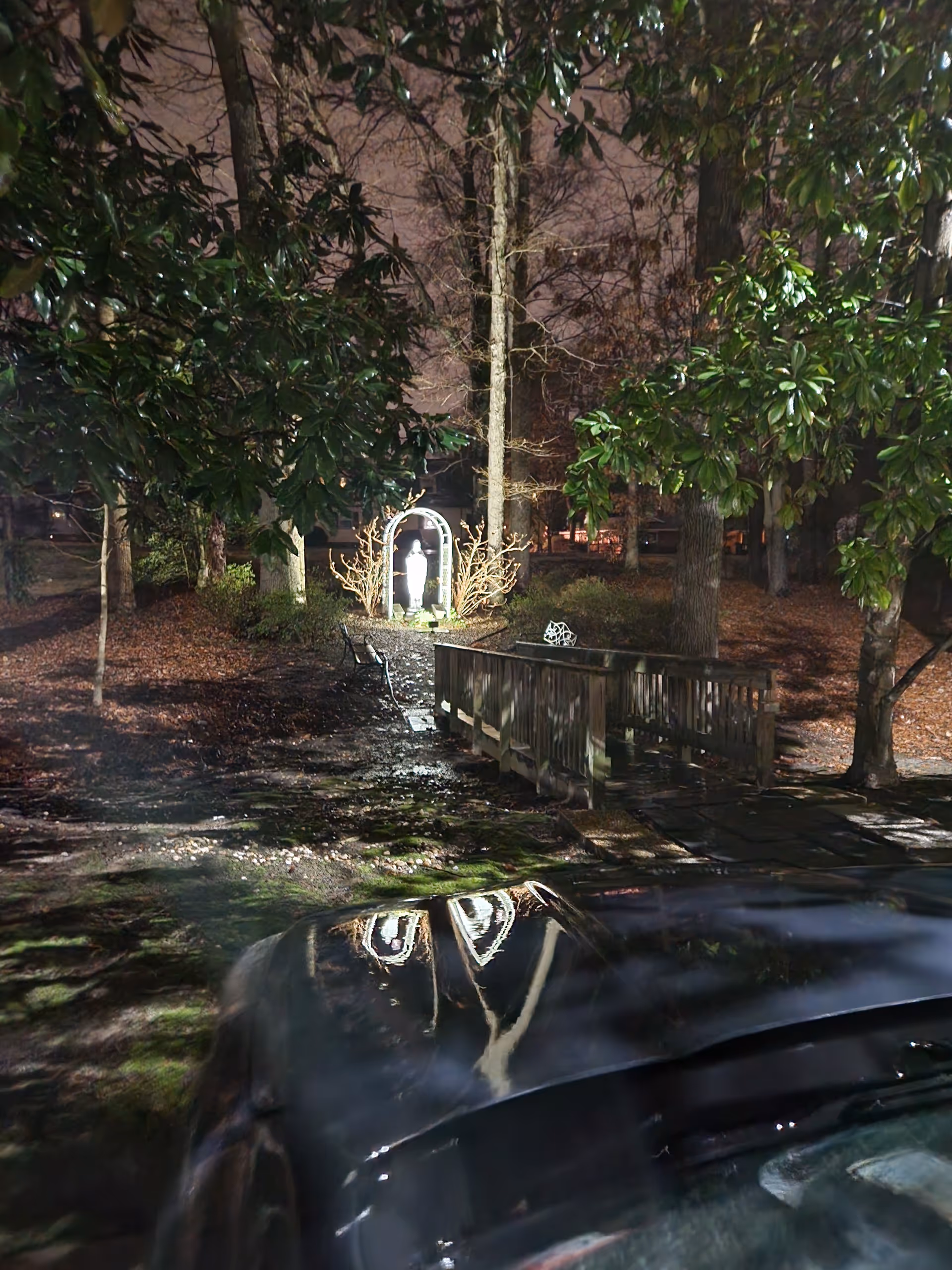 Nighttime view of a wooded outdoor area with a small statue, bridge, and car.