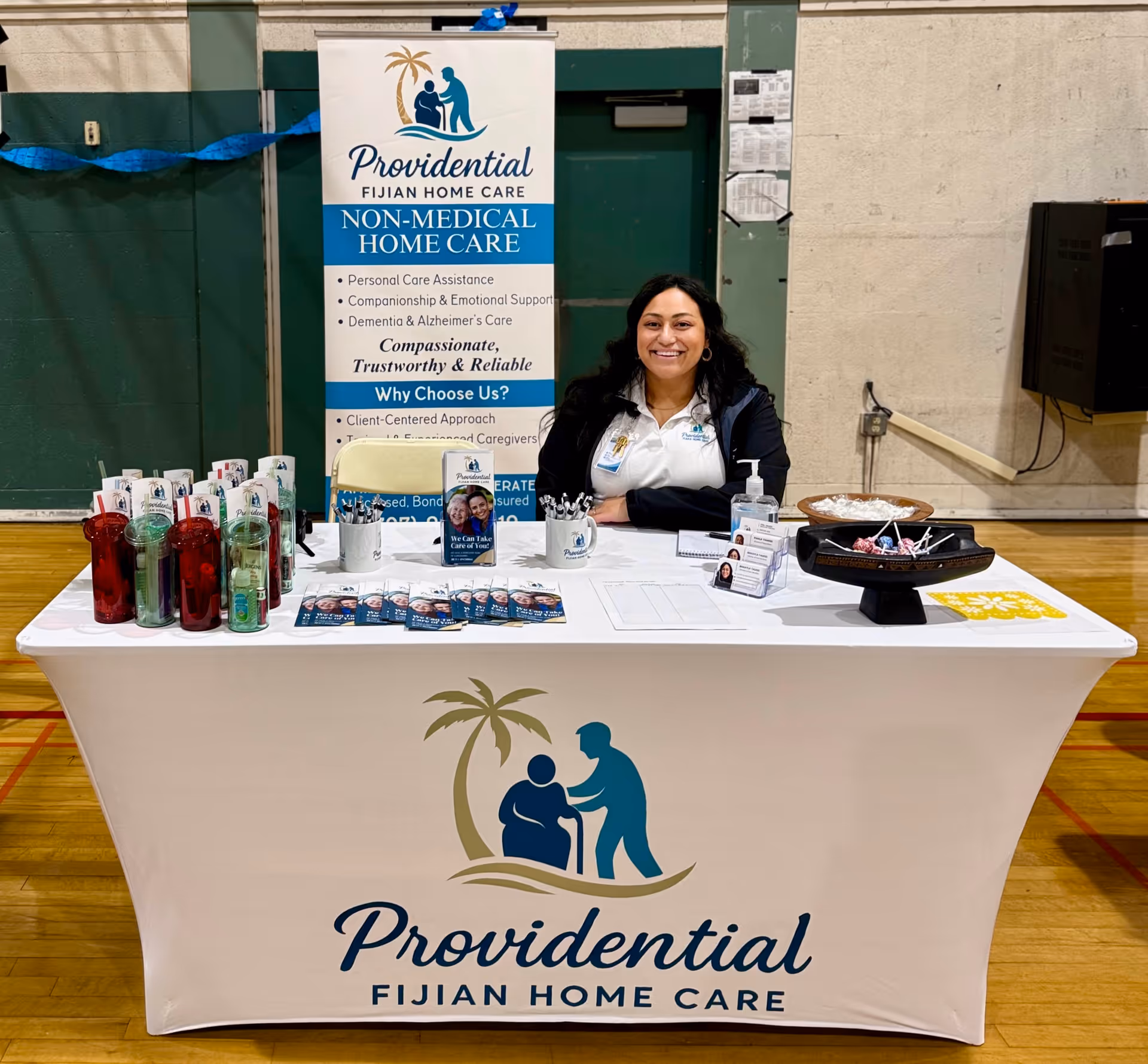Smiling woman sits at a branded information table promoting Providential Fijian Home Care in a gym.