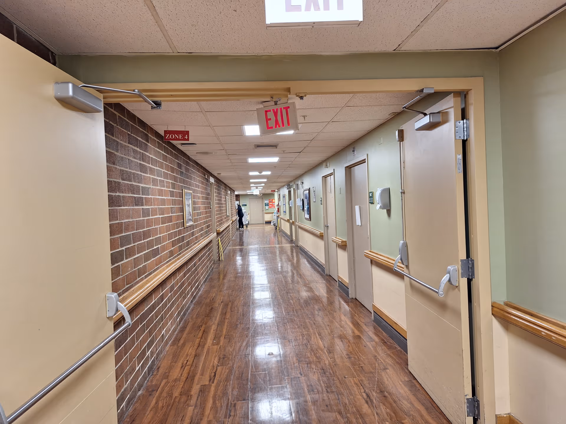 A long indoor hallway with wooden flooring and beige walls, featuring several closed doors on the right side and a brick wall on the left. There are handrails along both walls and an illuminated EXIT sign hanging from the ceiling. A person is visible in the distance at the end of the hallway.