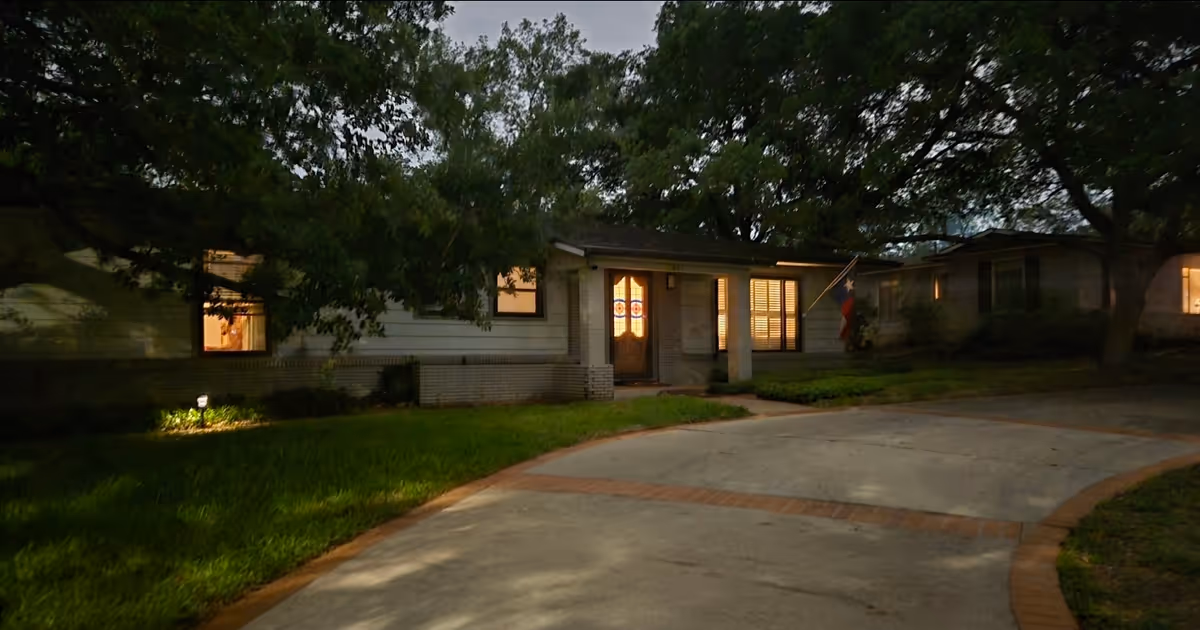 A single-story care home at dusk with lit windows and a visible Texas flag near entrance.