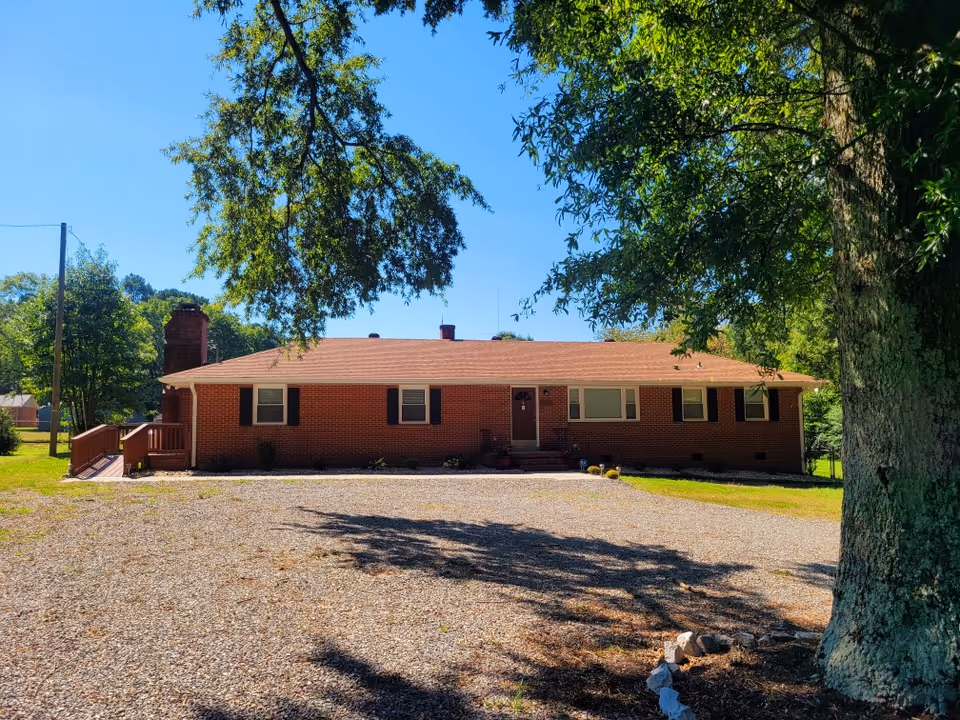 Single-story brick senior living facility front view with gravel driveway and large trees.