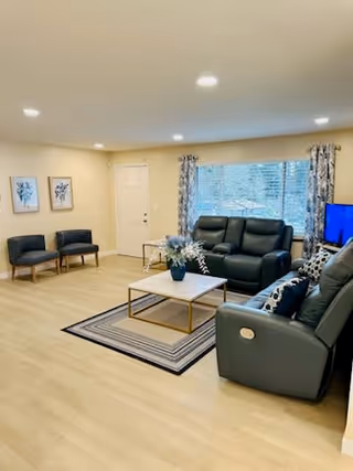 Well-lit living room featuring two reclining leather sofas, a coffee table on a striped rug, two chairs by the wall, and a TV near a large window.
