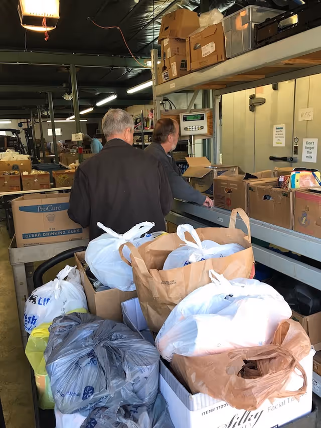 Two men stand by a conveyor in a cluttered supply room filled with cardboard boxes and plastic bags.