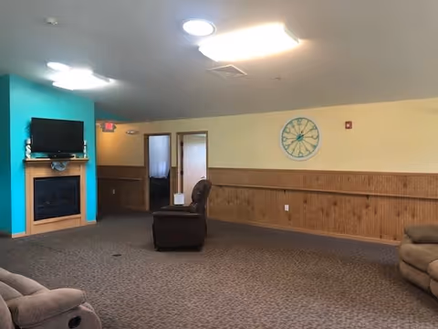 A common area in an assisted living facility with beige carpet, wood paneling on the walls, and a teal accent wall with a mounted TV above a fireplace. There are several recliner chairs arranged around the room and a large decorative clock on the wall.