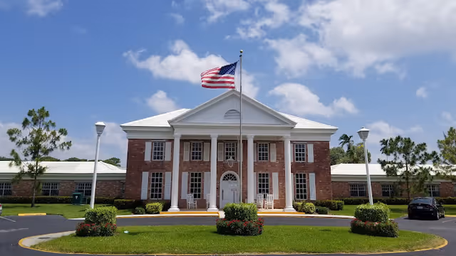 Front view of a red-brick, two-story building with white columns and an American flag on a flagpole.