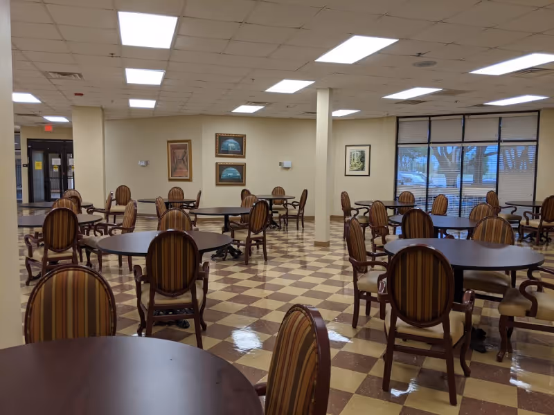 A spacious dining room with multiple round tables and striped cushioned chairs arranged neatly. The floor has a checkered pattern in beige and brown. The walls are light-colored with framed artwork hanging. Large windows with blinds allow natural light to enter the room.