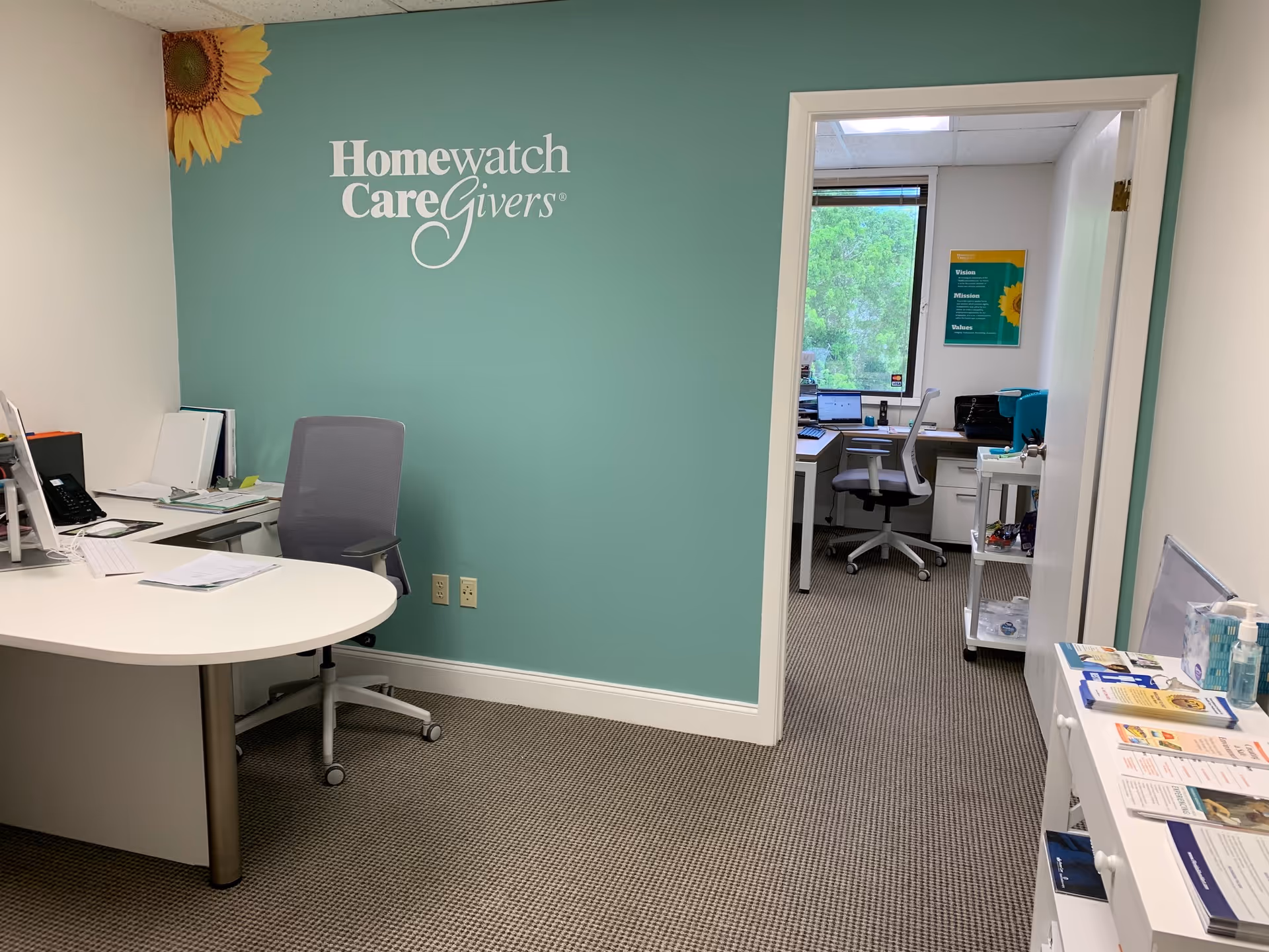 Office area with teal wall and Homewatch CareGivers logo, desks, chairs, and equipment.