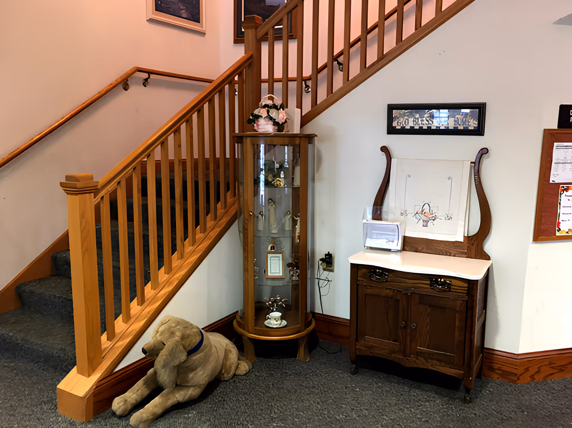 Carpeted stairwell interior with a wooden banister, glass curio cabinet, antique sideboard, and a large stuffed dog on the floor.
