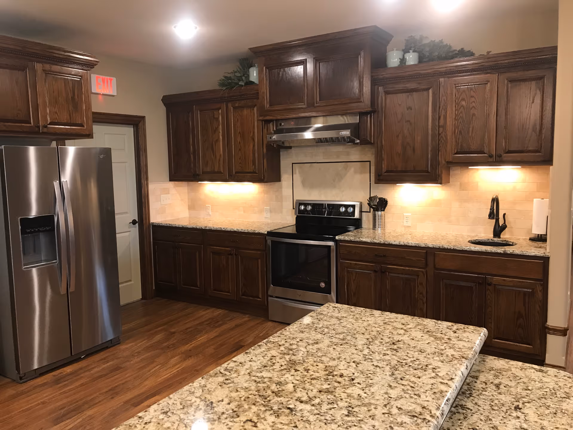 A kitchen with dark wooden cabinets, granite countertops, a stainless steel refrigerator, an electric stove with a range hood, a black faucet, and under-cabinet lighting. The floor is wooden, and there is an exit sign above a door in the background.