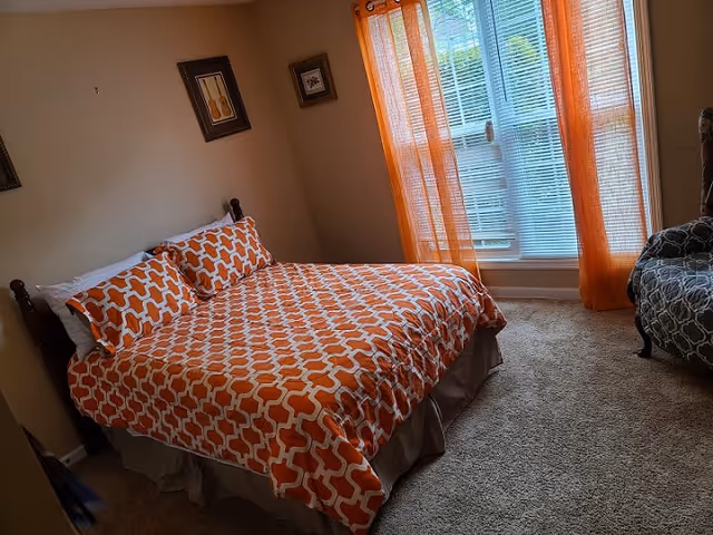 A bedroom with a bed covered in an orange and white patterned bedspread and matching pillows. There are two framed pictures on the beige wall above the bed. To the right, there is a window with orange sheer curtains and white blinds, and a patterned armchair is partially visible. The floor is carpeted in a light brown color.