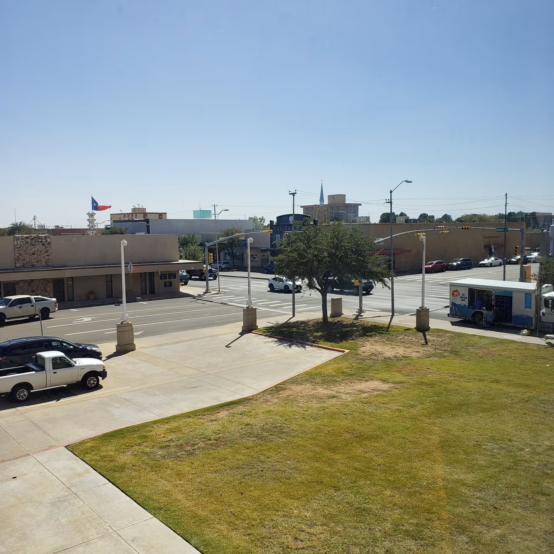 Wide outdoor street view with sidewalks, trees, and low-rise buildings on a sunny day.