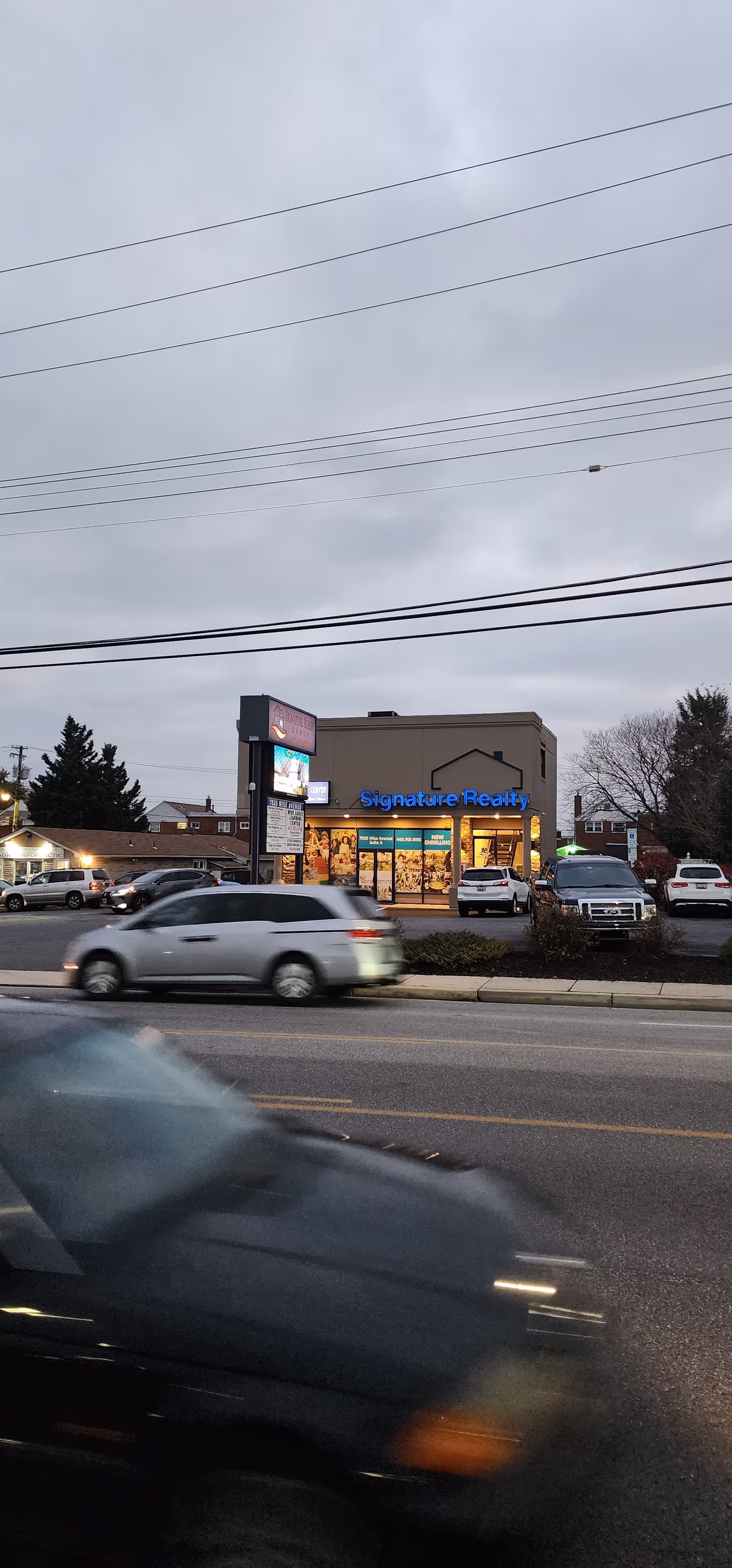 Front of a commercial building with Signature Realty sign and passing cars.