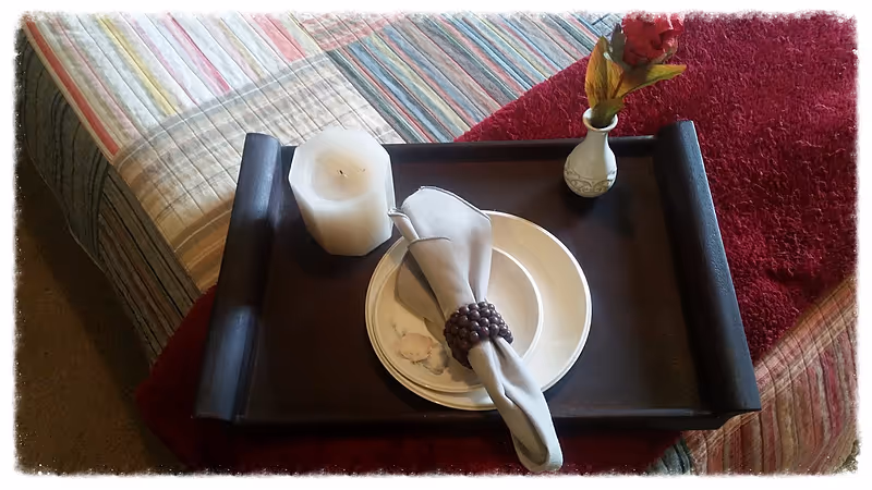 A black serving tray with a plate, napkin, candle, and small vase on a striped sofa.