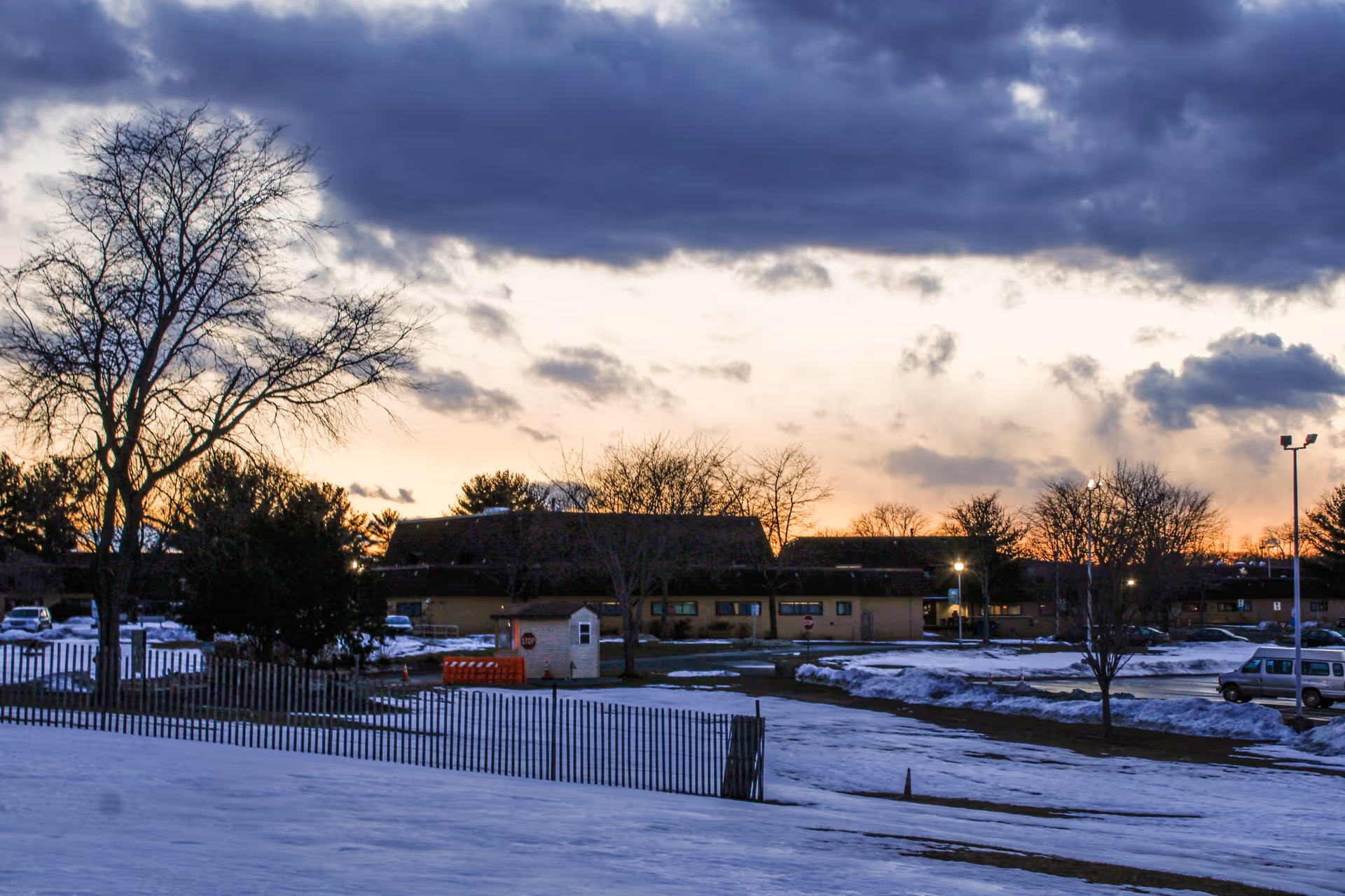 Snowy ground surrounds a low, long building at sunset, with trees and fence in foreground.