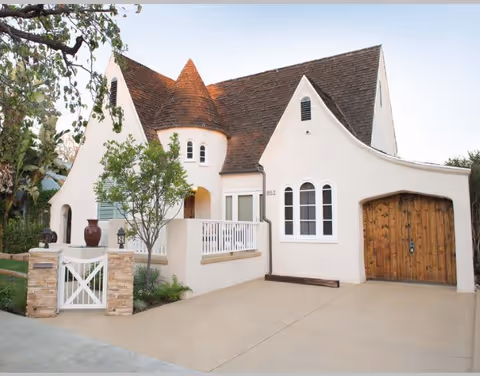 Front exterior of a white stucco cottage-style house with a turret, arched windows, small porch, and wooden garage door.