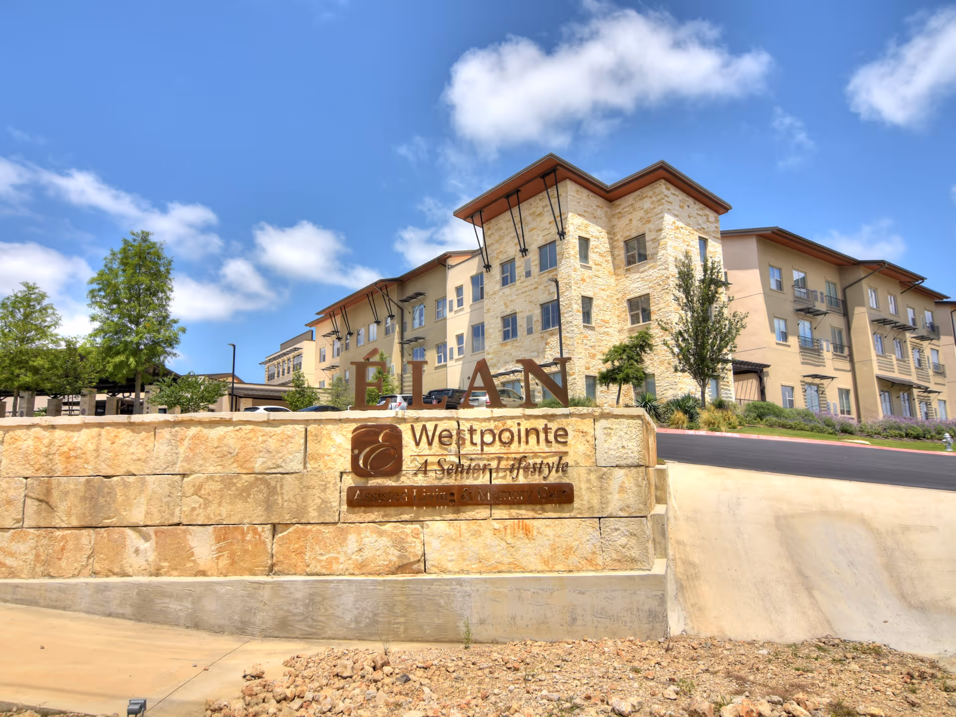 Exterior view of Élan Westpointe at New Braunfels senior living facility showing a multi-story building with stone and beige walls under a blue sky with some clouds. In the foreground, there is a stone sign with the facility name and logo.