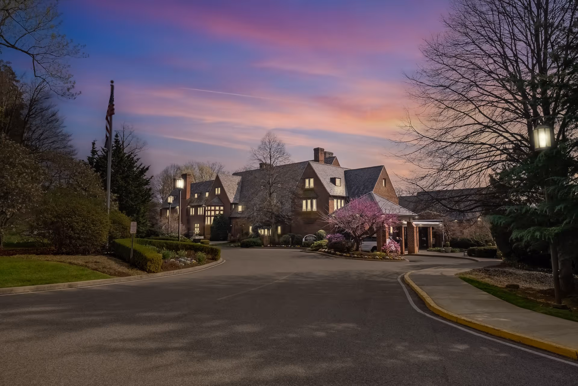 Exterior view of The Quadrangle facility at dusk with a large brick building, illuminated windows, a driveway, landscaped bushes, trees, and a flagpole under a colorful sky.