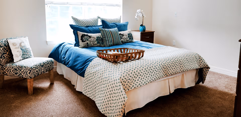 A cozy bedroom with a neatly made bed featuring blue and white bedding and multiple decorative pillows. There is a wooden tray on the bed, a patterned upholstered chair with a cushion to the left, a wooden nightstand with a blue vase and white flowers, and a window with sheer curtains letting in natural light.