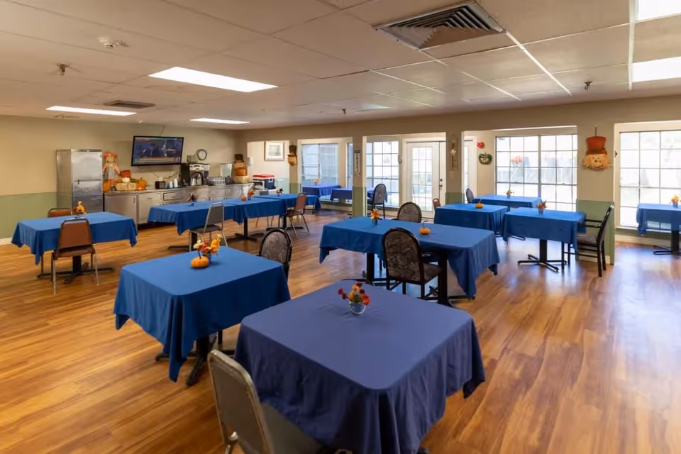 Spacious dining room with several square tables draped in blue tablecloths, chairs, small autumn centerpieces, and a serving counter along the back wall.