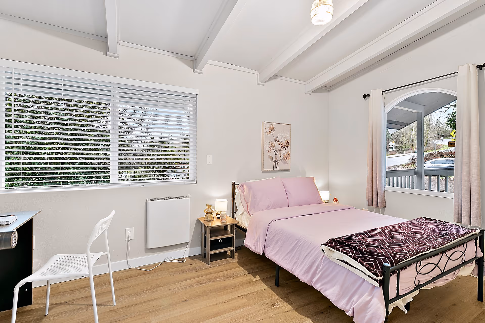 A bright bedroom with pink bedding, a desk and white chair, and large window blinds.