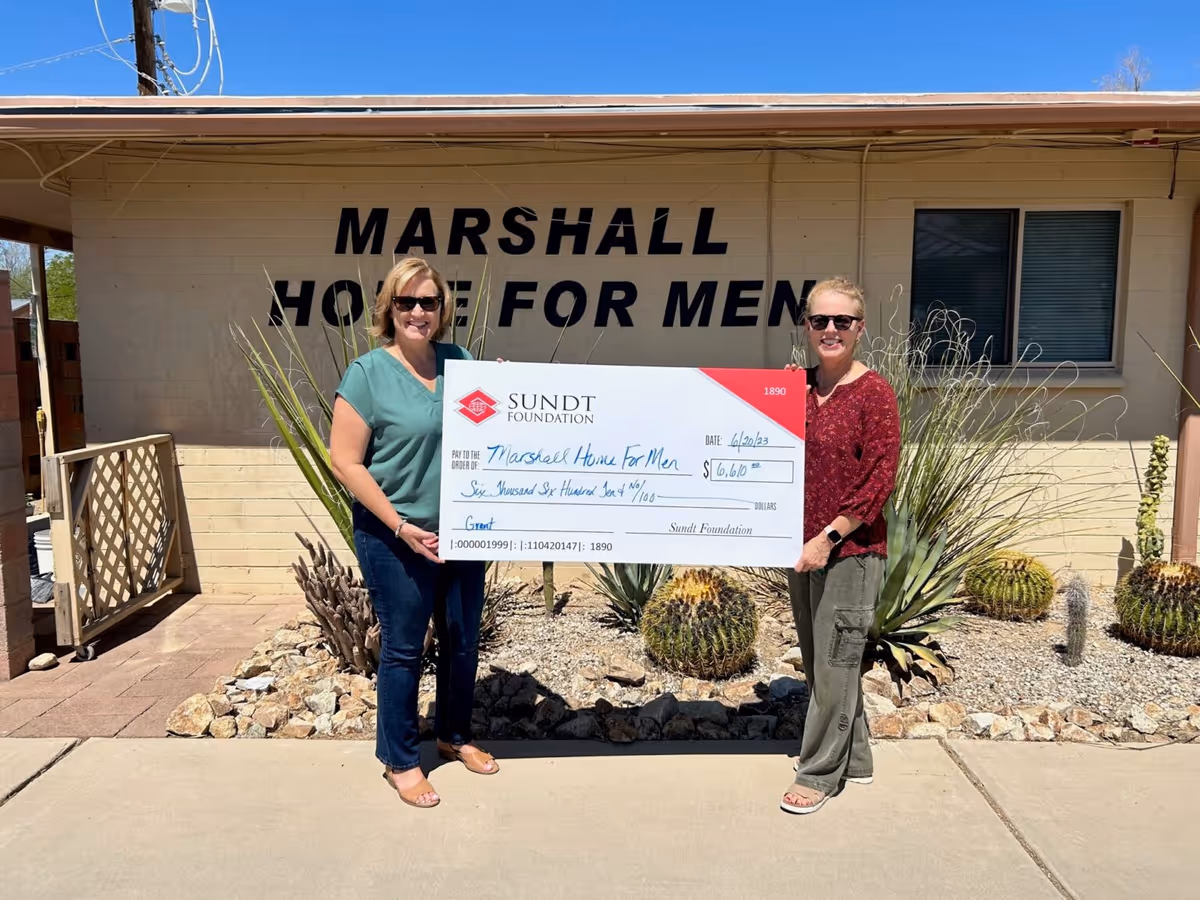 Two women standing outside a building labeled "Marshall Home for Men" holding a large ceremonial check from the Sundt Foundation.