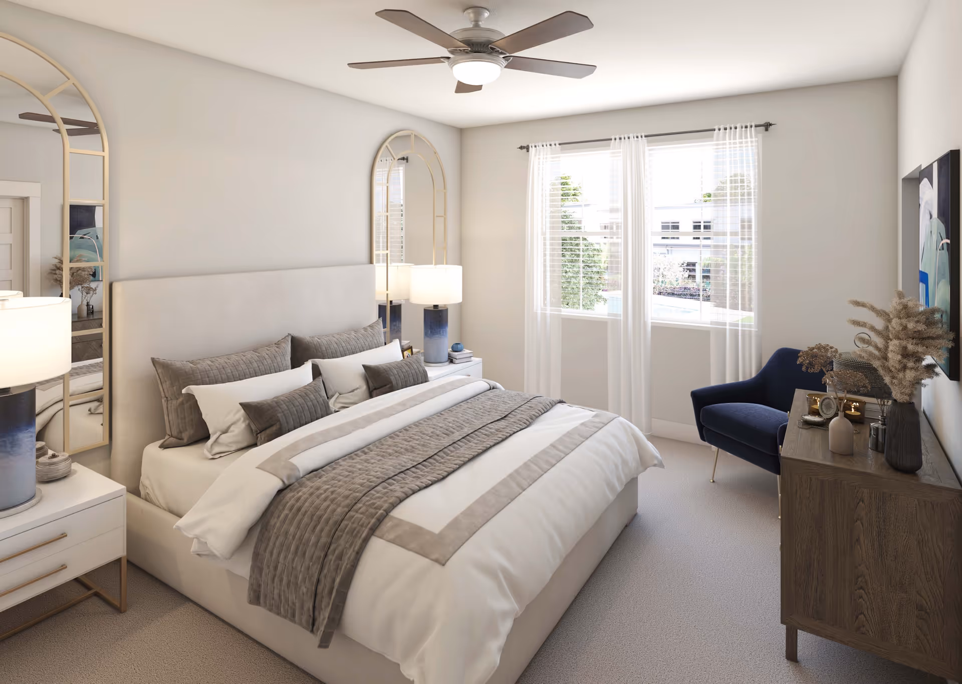 A bright and cozy bedroom featuring a large bed with white and gray bedding, two bedside tables with lamps, a ceiling fan, a blue armchair, a wooden dresser with decorative items, and two tall arched mirrors on the wall. A window with sheer white curtains lets in natural light.
