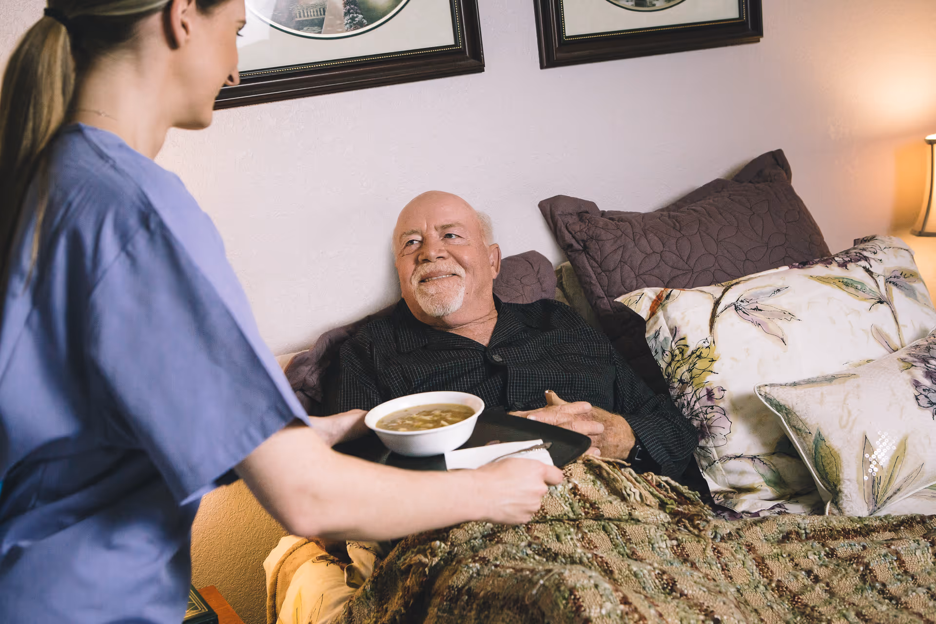 Caregiver in blue scrubs feeds soup to elderly man in bed.