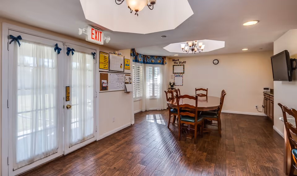 Bright communal dining room with a wooden round table and chairs, French doors with an EXIT sign, and a wall-mounted TV.