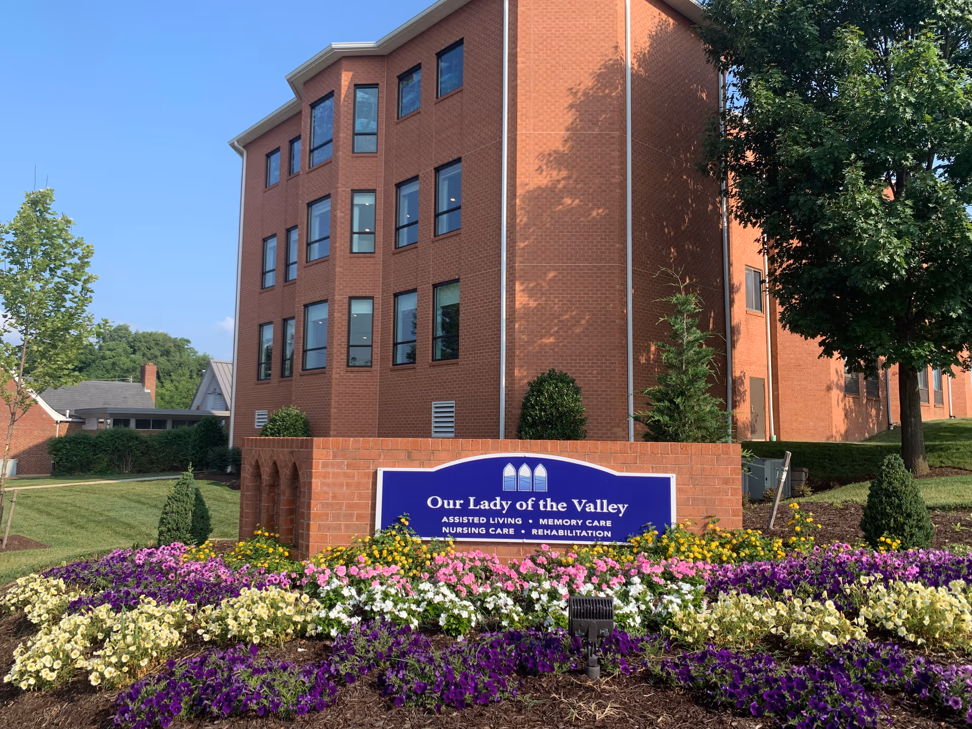 Exterior view of a multi-story brick building with multiple windows, surrounded by well-maintained landscaping including colorful flower beds and green trees. In front of the building is a blue sign that reads 'Our Lady of the Valley Assisted Living, Memory Care, Nursing Care, Rehabilitation.'