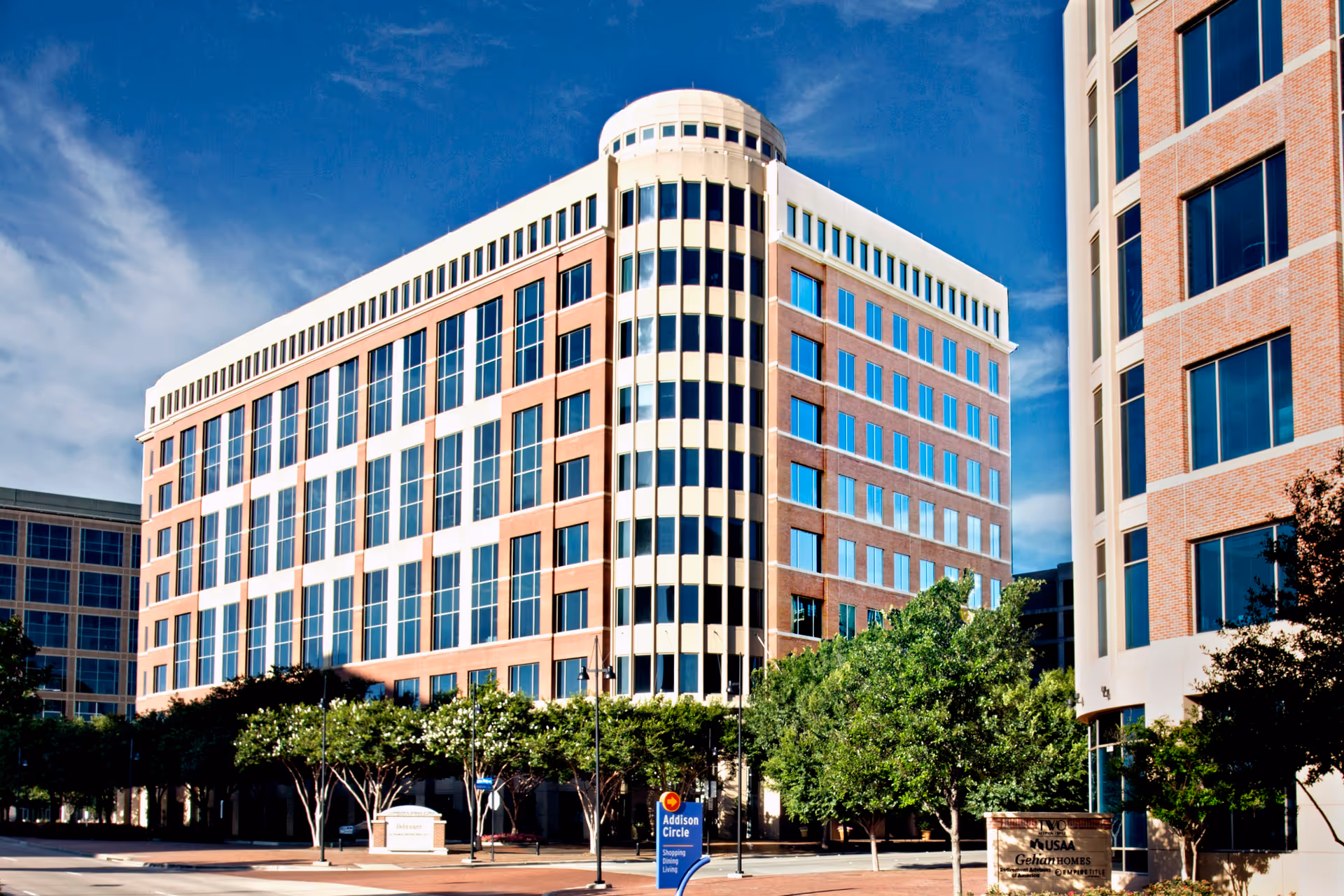 A multi-story brick building with glass windows and surrounding trees under a blue sky.