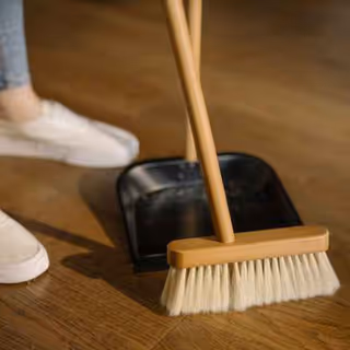 A broom and dustpan resting on a wooden floor beside a person's sneakers.