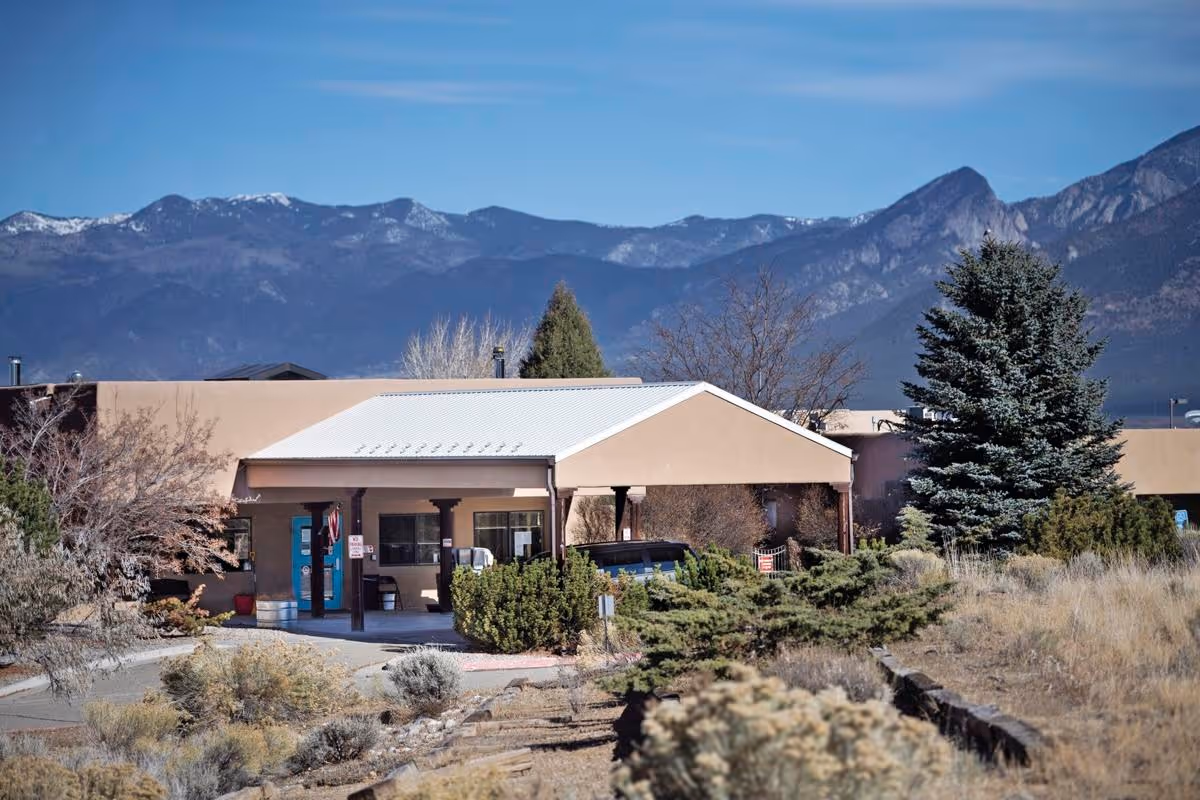 Exterior view of Taos Healthcare facility with a beige building, a covered entrance, surrounded by shrubs and trees, and mountains in the background under a clear blue sky.