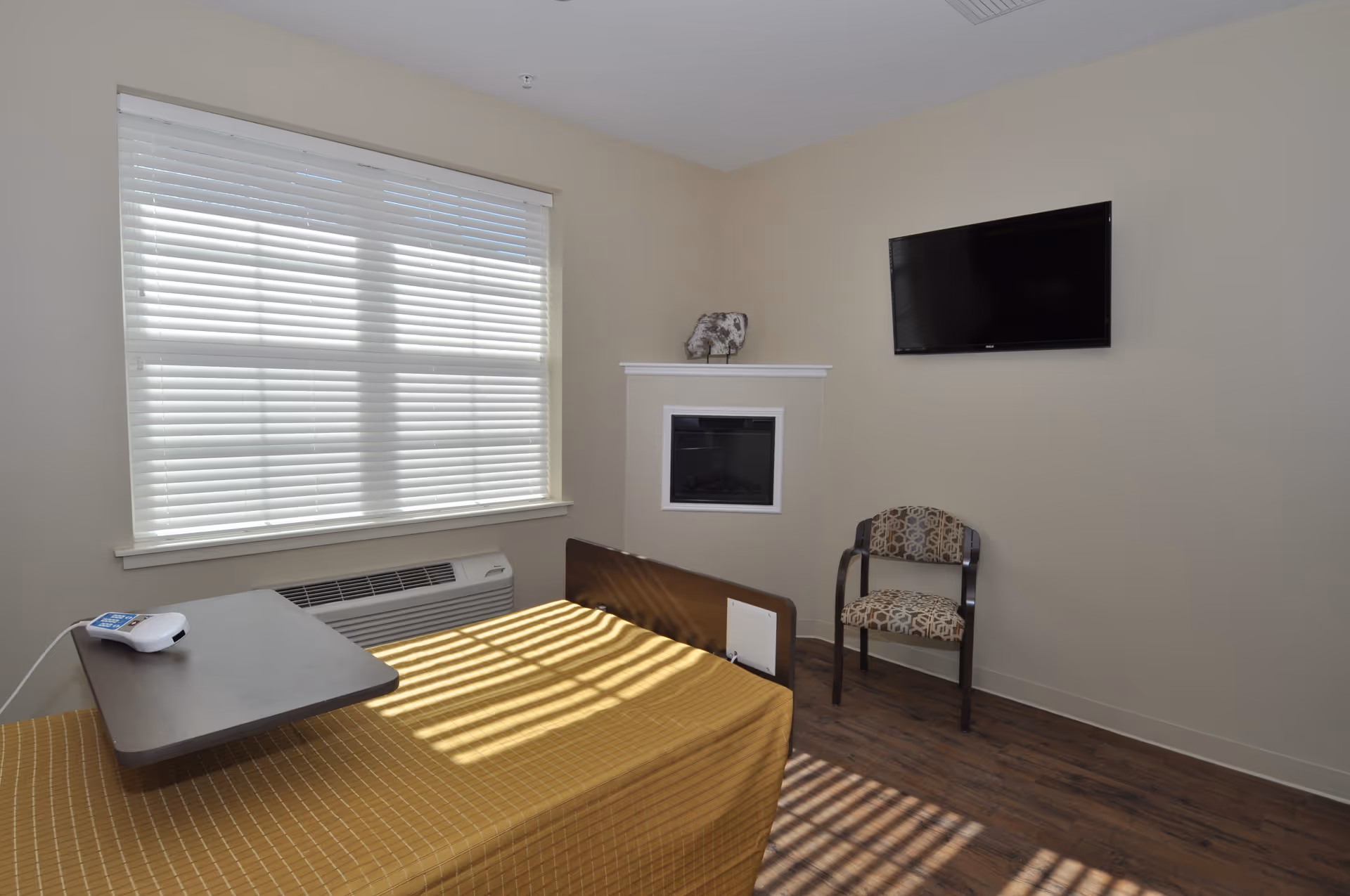 Sunlit bedroom with bed, blinds, fireplace, chair, and wall-mounted TV.