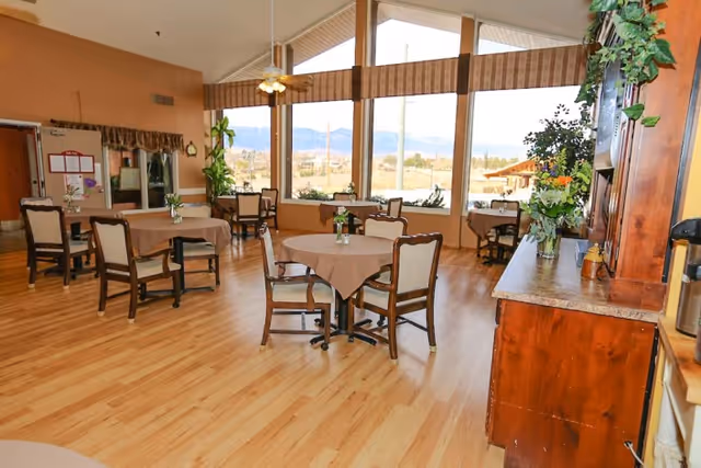 A bright dining room with wooden floors and several round tables covered with beige tablecloths, each surrounded by wooden chairs with cushioned seats. Large windows provide a scenic view of mountains and open land outside. There are plants and flowers decorating the room, and a wooden cabinet with more plants and a coffee maker is visible on the right side.