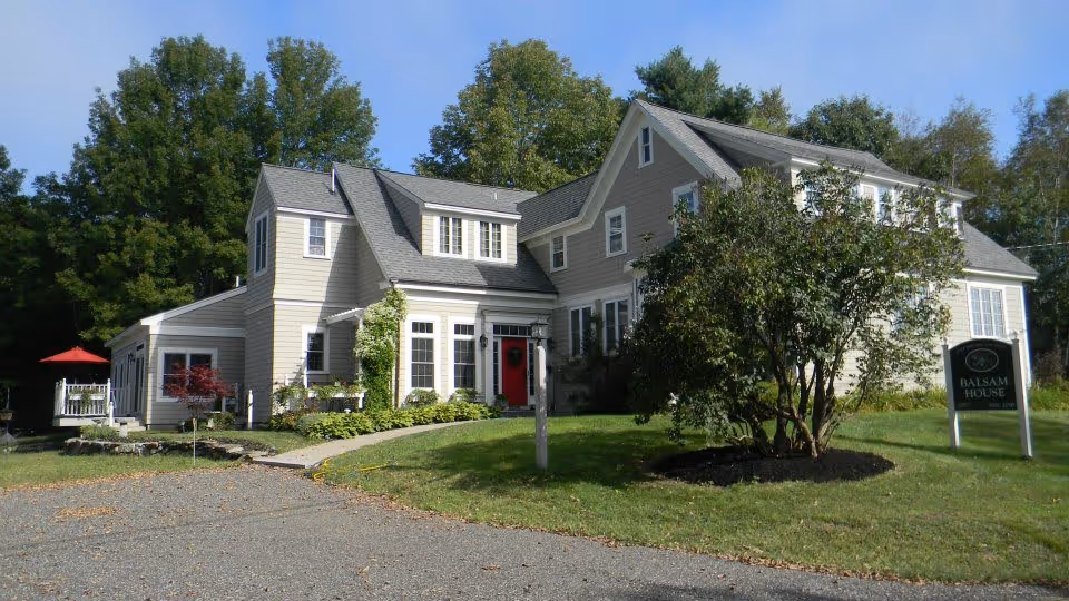 Exterior view of a large, light gray two-story house with a red front door, surrounded by green trees and a well-maintained lawn. There is a sign on the right side of the image that reads 'Balsam House'. A red umbrella is visible on a patio area to the left.