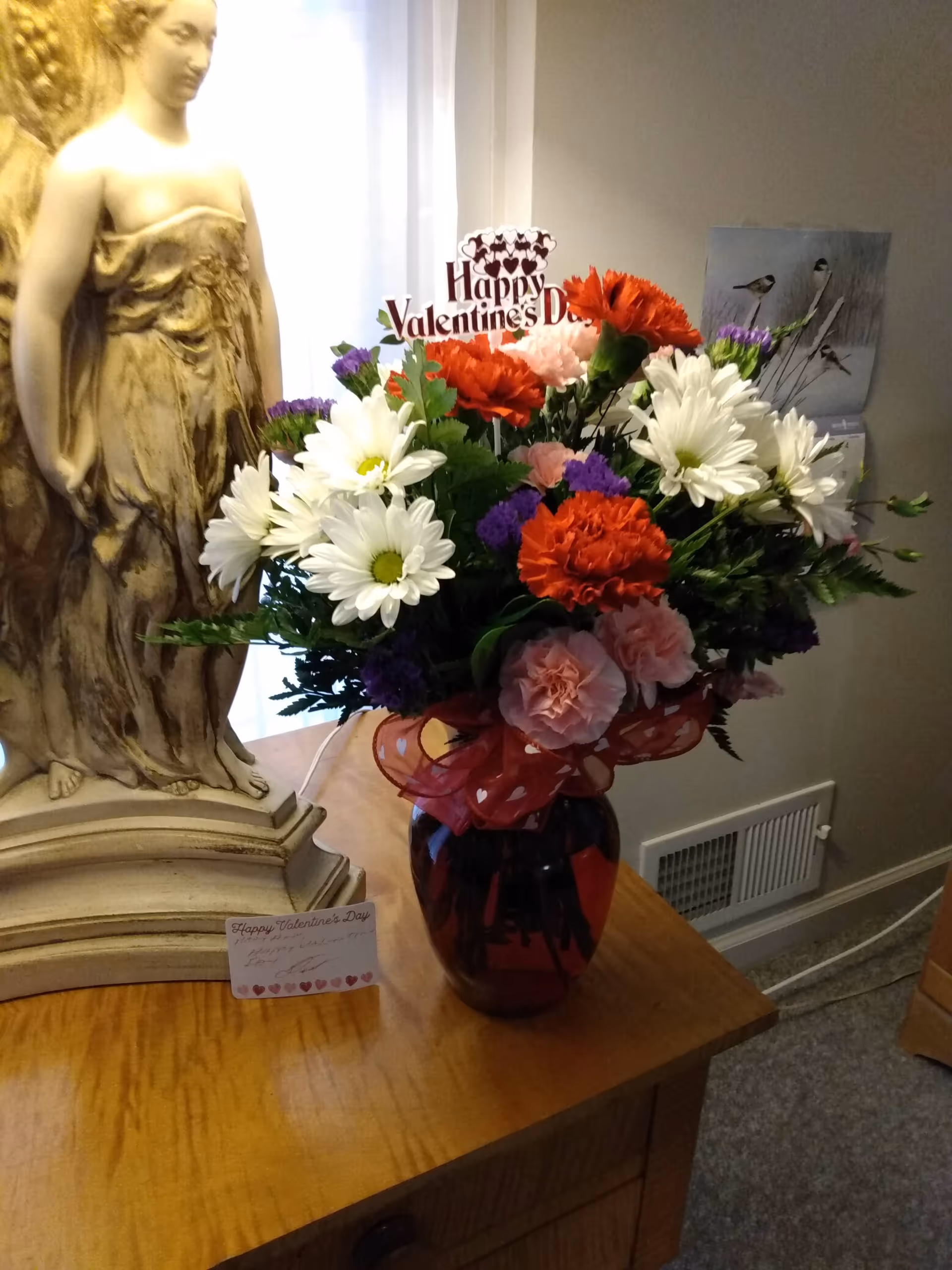 Statue on a wooden table with a vibrant flower arrangement and Valentine's Day sign indoors.