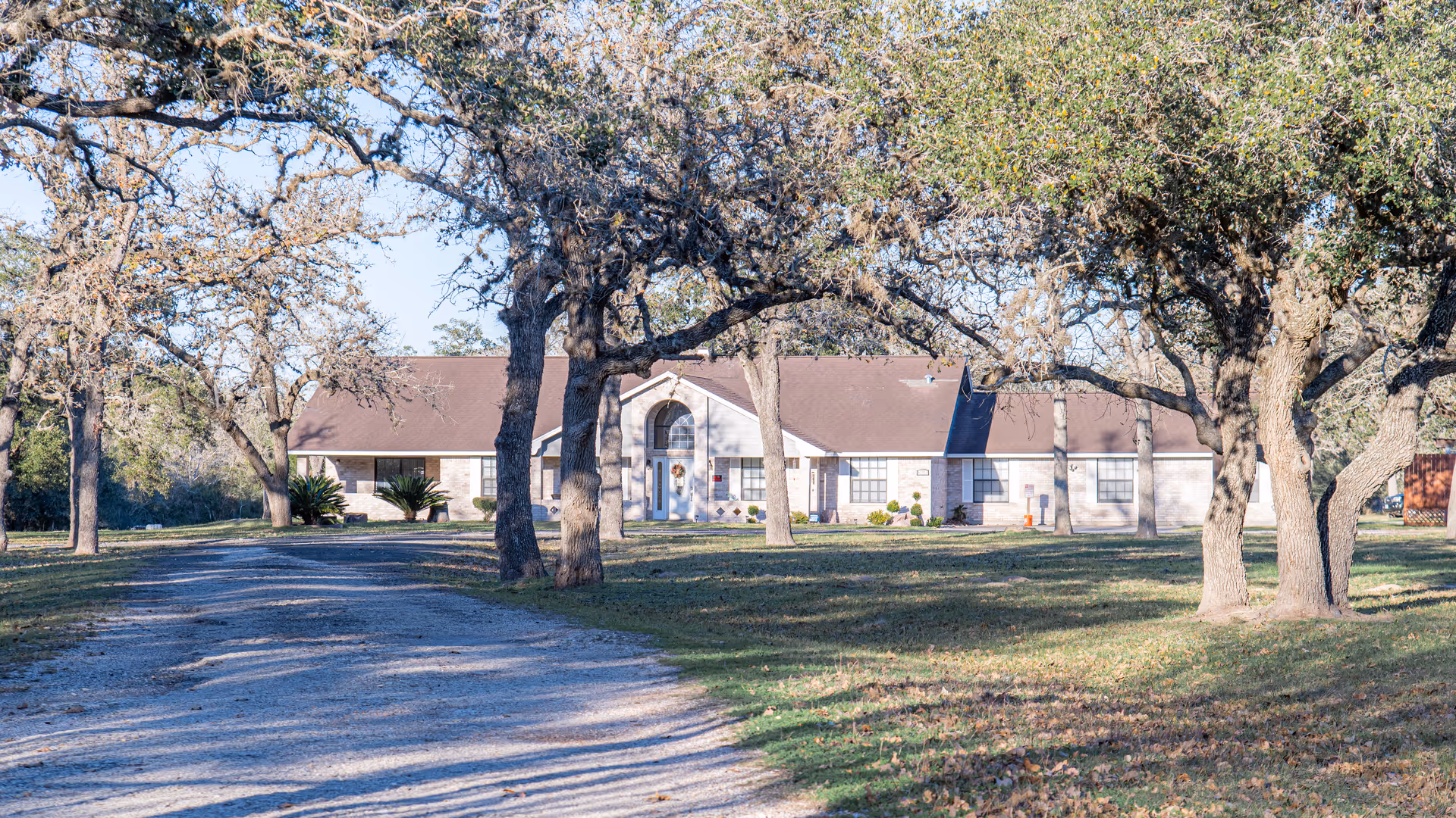 Single-story assisted living building with a gravel driveway, trees, and an arched front entrance.