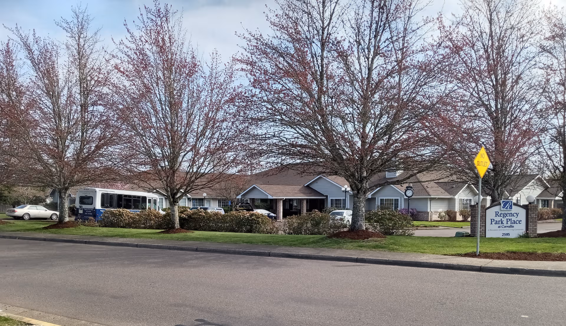 Front view of Regency Park Place at Corvallis with trees, sign, a bus, and cars
