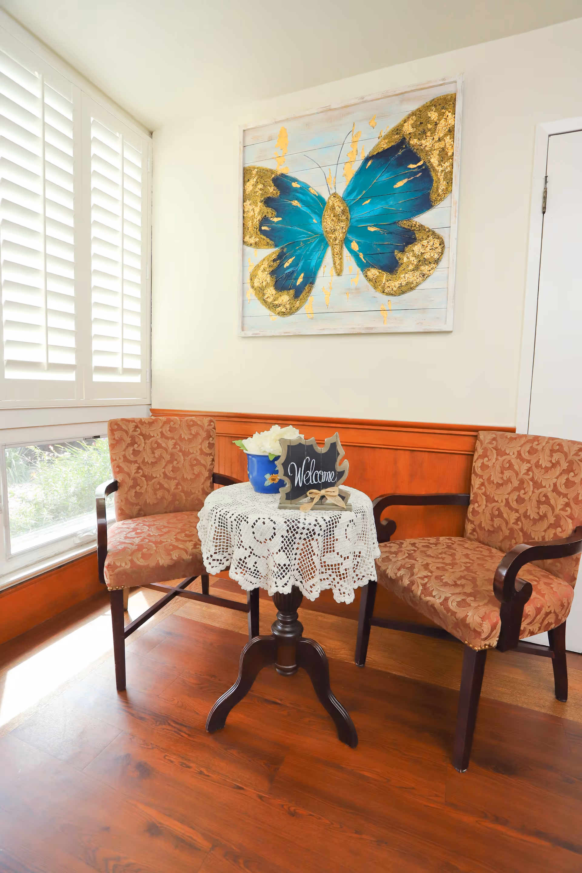 Two patterned chairs surround a lace-covered round table in a lobby seating area, with butterfly art.