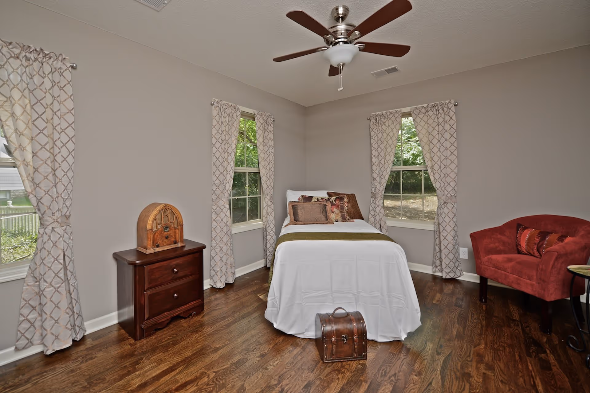A bedroom with a single bed covered in white bedding and decorative pillows, positioned between two windows with patterned curtains. There is a dark wooden nightstand with a vintage radio on it to the left of the bed, and a red upholstered armchair with a decorative pillow to the right. The room has wooden flooring and a ceiling fan with wooden blades.