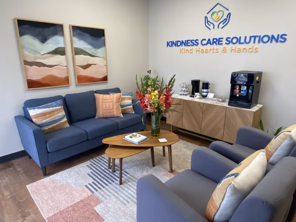 A welcoming lounge with blue sofas, coffee tables, and a coffee station beside Kindness Care sign.