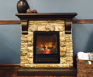 A small stone electric fireplace with a dark wood mantel against a blue wall, a decorative vase on top and a wicker basket to the right.