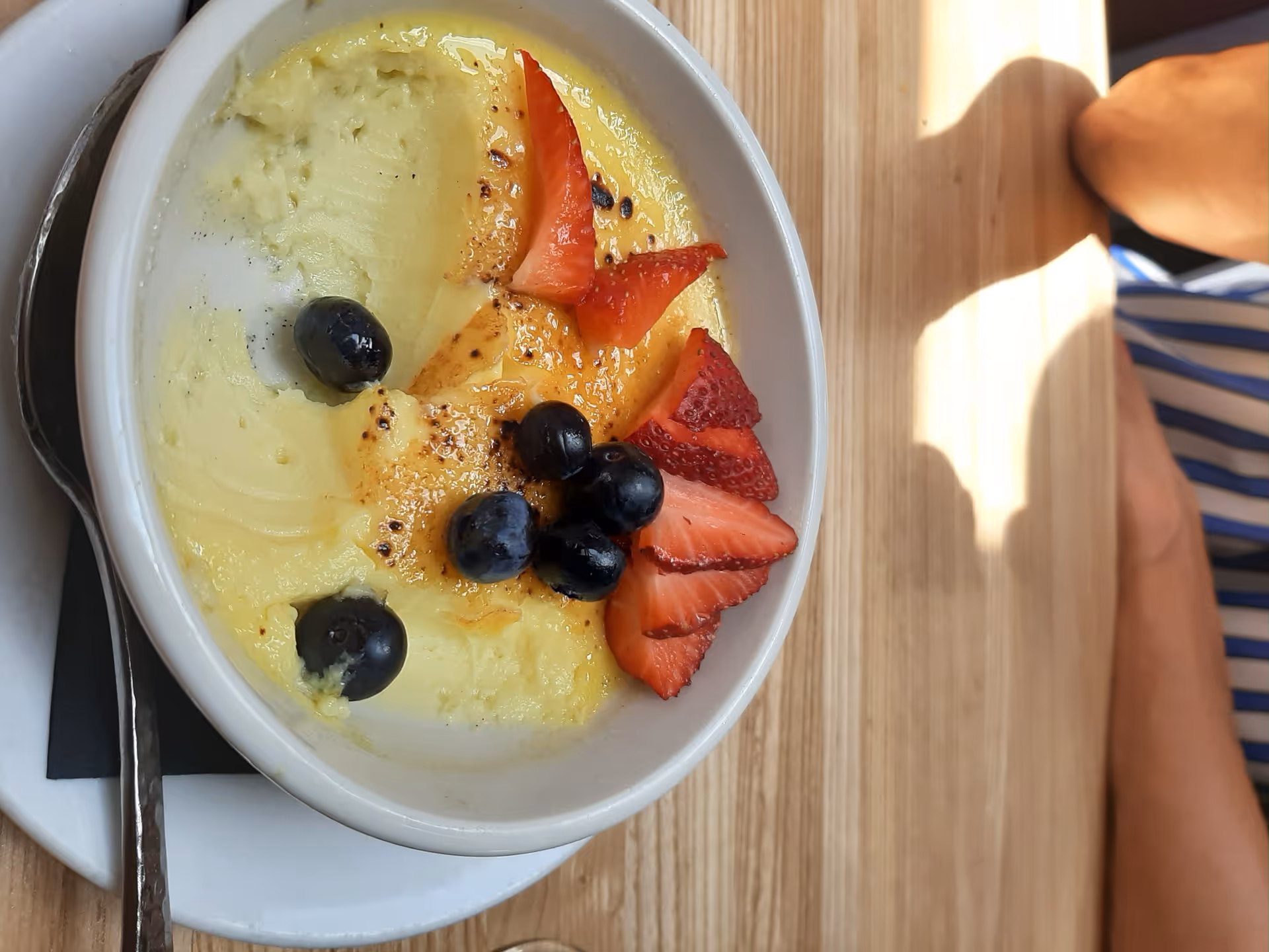 A bowl of custard or pudding topped with sliced strawberries and blueberries on a wooden table, with a spoon resting on a napkin beside the bowl. A person's arm wearing a blue and white striped shirt is partially visible on the right side.
