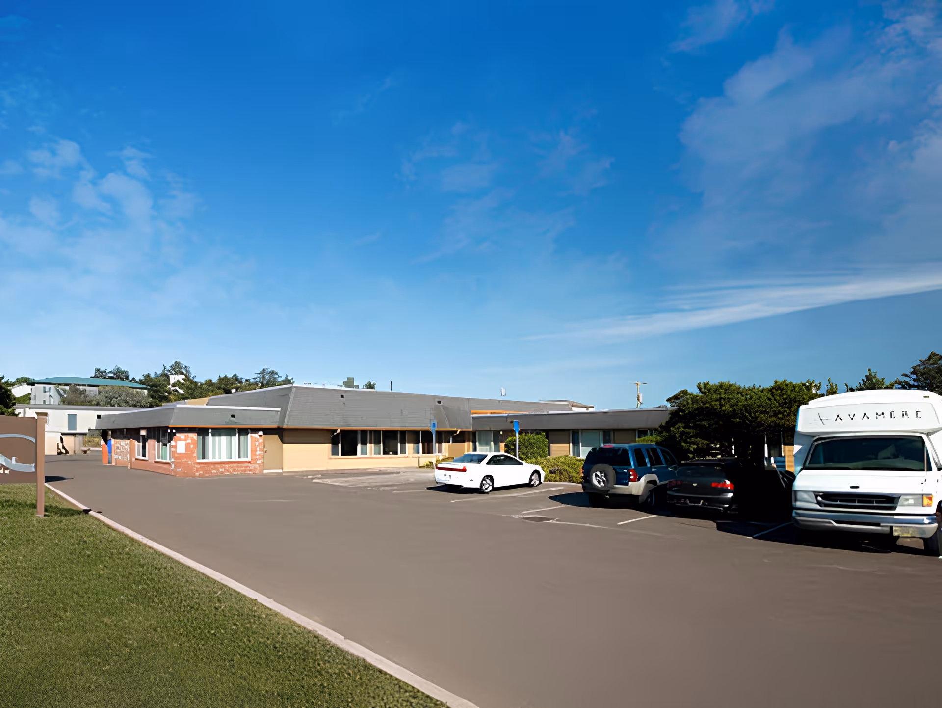 Single-story rehabilitation facility building with a parking lot and several parked vehicles under a clear blue sky.
