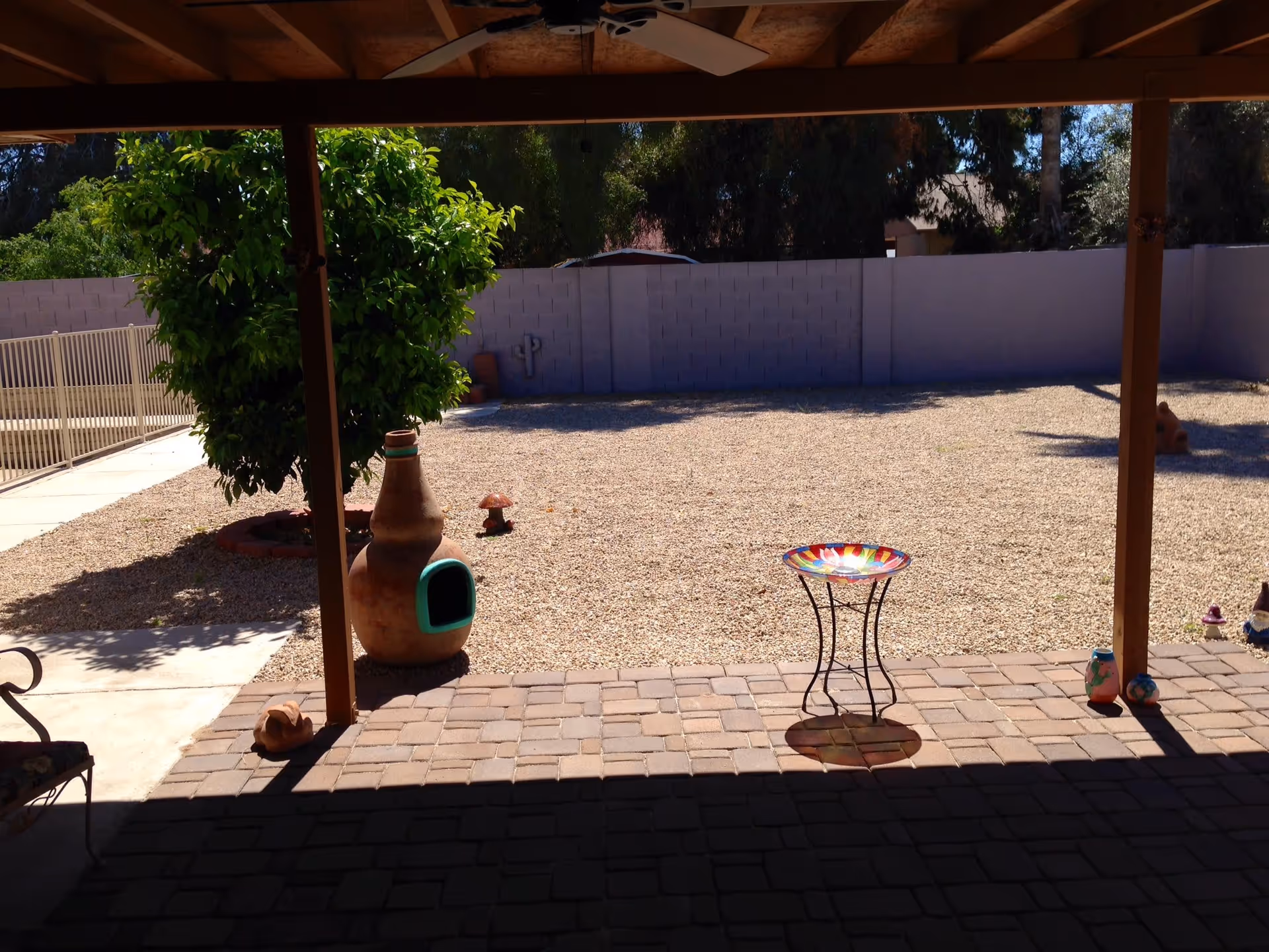Covered patio looking out onto a gravel backyard with a tree, chiminea, and a colorful birdbath on a paver patio.