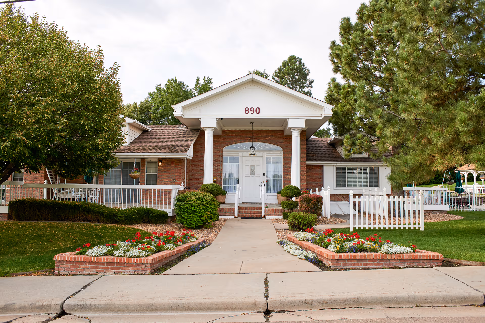 Brick senior living facility front with white columns, landscaped flower beds, and a walkway.