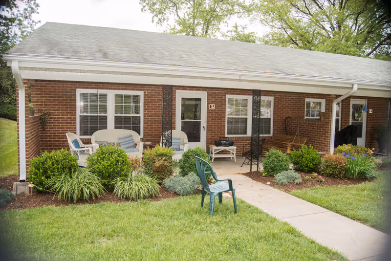 Front view of a single-story brick building with a covered porch area featuring outdoor seating including white wicker chairs, a wooden swing, and a green plastic chair on the lawn. The porch is surrounded by neatly trimmed bushes and plants, with a concrete walkway leading to the entrance doors.