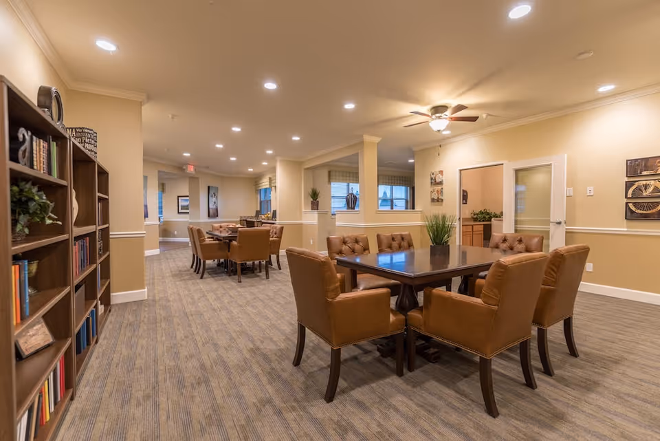 Well-lit senior living common area with a large dining table and tan leather chairs.