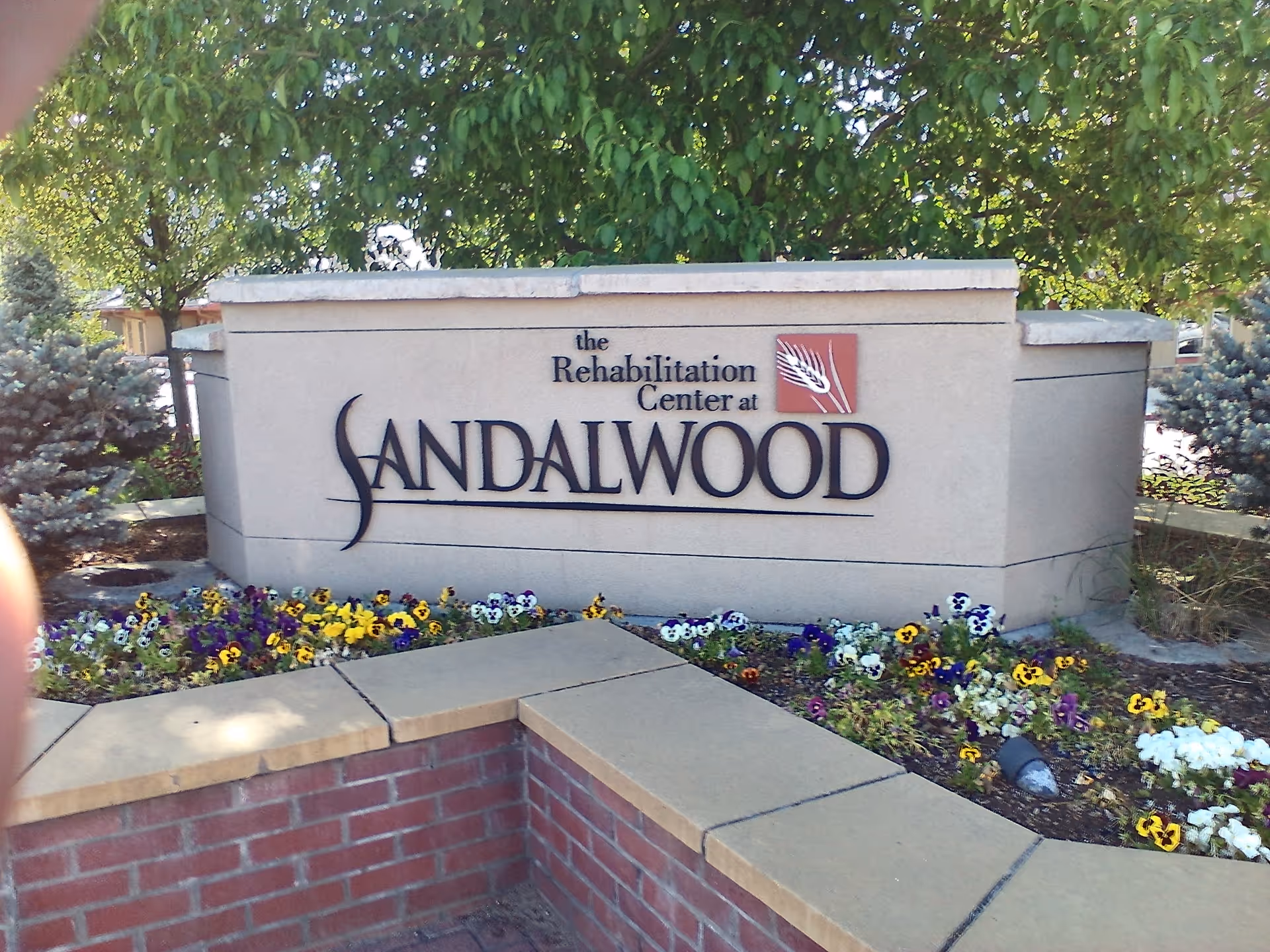 Rehabilitation Center at Sandalwood sign displayed outdoors with flowerbeds and trees.