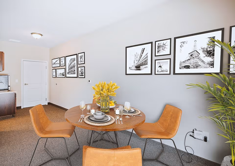 Bright interior dining area with a round wooden table set for four, tan leather chairs, framed black-and-white wall art, and a kitchenette in the background.
